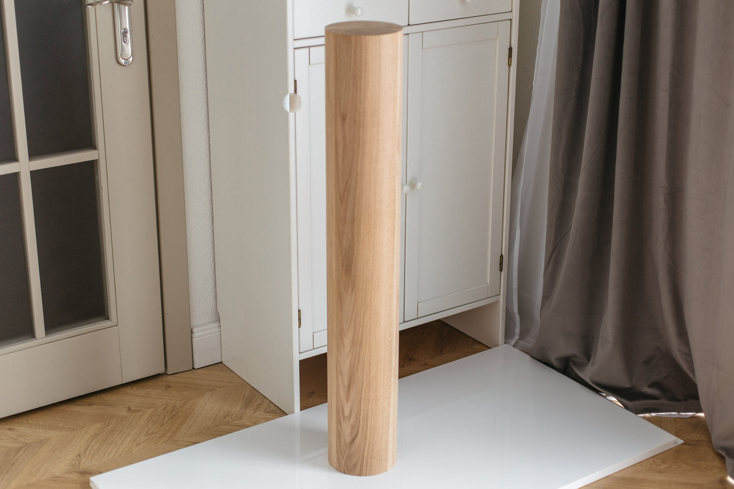 A tall, cylindrical wooden column or lamp base with a natural oak wood grain finish, standing upright on a white surface in a room setting with a white cabinet, gray curtain, and wooden floor.